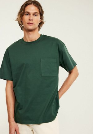 Junger Mann mit welligem Haar, der ein dunkelgrünes T-Shirt mit kurzen Ärmeln und einer Brusttasche trägt, vor einem einfarbigen hellen Hintergrund stehend.