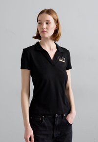 Polo preto com colarinho, mangas curtas e logo "EA7" dourado no peito. Tecido leve com uma textura suave.