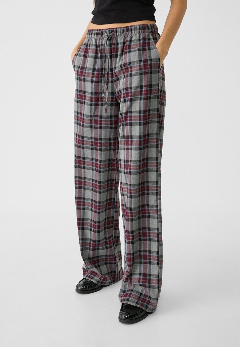 Osoba w luźnych, szarych i czerwonych spodniach w kratę z elastycznym paskiem i sznurkiem, z rękami w kieszeniach, w czarnych butach na białym tle.