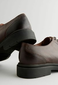 Chaussures en cuir marron avec une finition texturée, dotées d'une semelle en caoutchouc noir et de bouts arrondis. Les lacets et les coutures sont visibles.