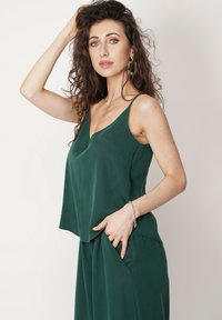 Zielona, bezrękawnikowa bluzka o luźnym kroju, dekolcie w literę V i miękkiej fakturze. Modelka pozuje z ręką w kieszeni, prezentując swobodny styl.