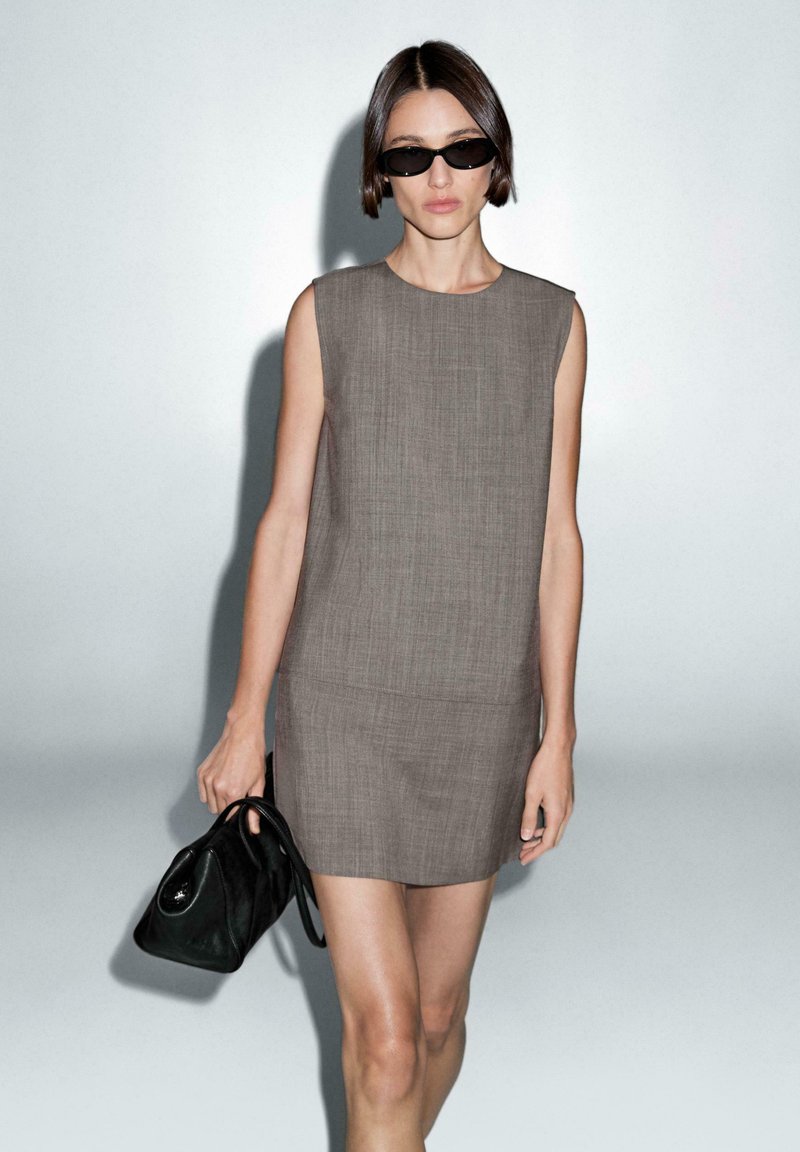 Gray, sleeveless mini dress with a textured design. Model holds a black bag, wearing dark sunglasses, against a neutral background.