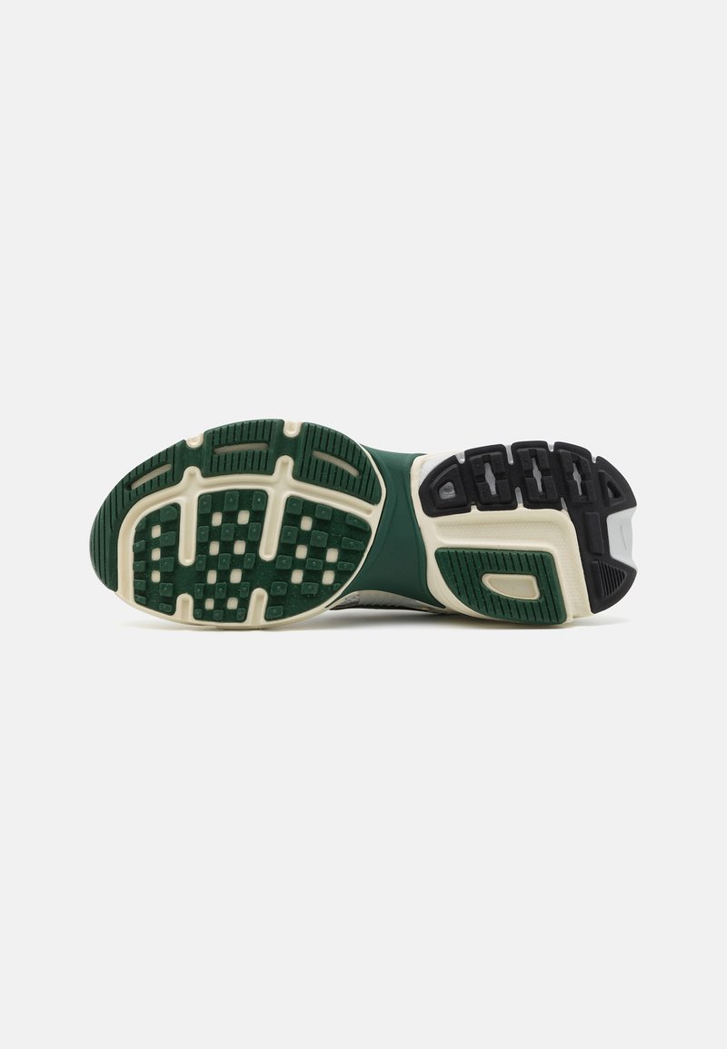 Suela de unas zapatillas en verde, blanco y negro con patrones de agarre segmentados y textura, presentada sobre un fondo blanco liso.
