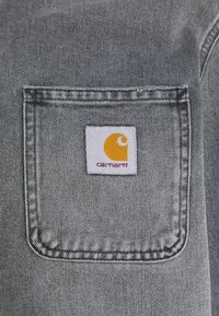Tejido de mezclilla gris con un bolsillo rectangular que lleva un parche con el logo de Carhartt en amarillo y blanco cosido en el centro.