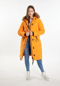 Dlouhý oranžový padded kabát s kapucí podšitou kožešinou, černými akcenty, zipovým zapínáním a šňůrkami. Nosený přes džíny a vícolorové tenisky.