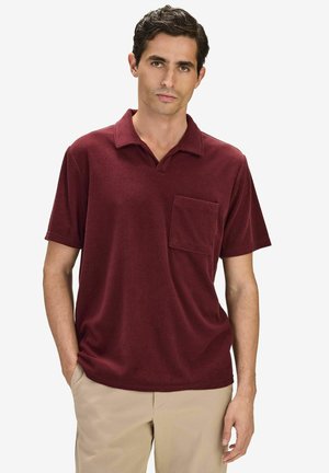 Mann trägt ein kurzärmeliges burgunderrotes Poloshirt mit Brusttasche und beigefarbene Hose, steht mit einer Hand in der Tasche.