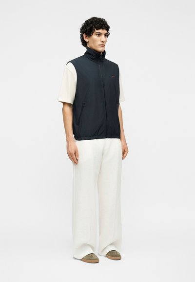 Άνδρας με σγουρά μαλλιά, που φοράει μαύρο γιλέκο με φερμουάρ πάνω από λευκό κοντομάνικο πουκάμισο, λευκό παντελόνι φαρδύ στα πόδια και ελιά πράσινα παπούτσια, στεκόμενος και κοιτάζοντας δεξιά.