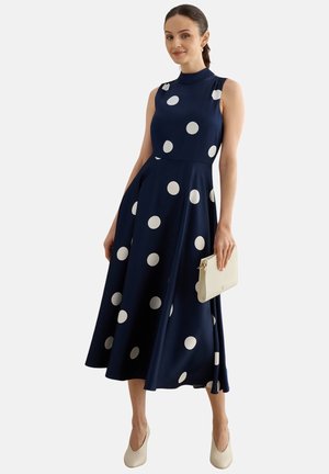 Frau trägt ein ärmelloses marineblaues Kleid mit großen weißen Punkten, hält eine weiße Clutch und trägt cremefarbene Absätze.