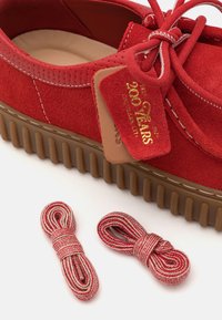Zapatos de ante rojo con una gruesa suela de goma marrón. Presentan un lazo y una etiqueta que dice "200 años". Incluye dos pares de cordones rojos y blancos.
