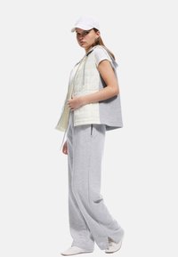 Leichte daunenartige Weste mit grauen Akzenten, getragen über einem grauen Shirt und Jogginghosen. Eine weiße Mütze und Sneaker vervollständigen den Look.