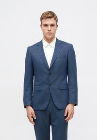 Sacou bleu, croit cu un rever cu șanț, cu un singur rând de nasturi, două nasturi, și două buzunare frontale, purtat cu o cămașă albă.