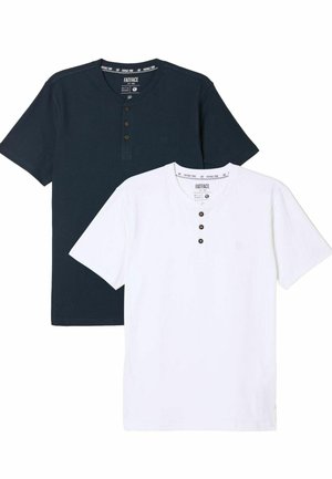 Smilende mand med skæg står med armene over kors iført navy kortærmet henley-trøje, mørkeblå jeans og olivengrønne sneakers på gråt gulv.