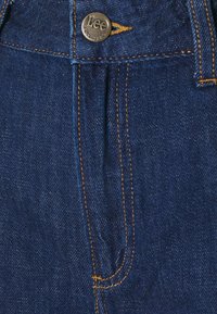 Fâlcul cu nasturi al unei perechi de blugi din denim albastru cu cusături galbene și un nasture metalic gravat cu "Lee Est. 1889."