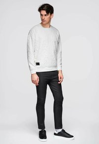 Homme debout, portant un sweat-shirt blanc embossé, un jean noir slim, des baskets noires à semelles blanches, sur un fond gris clair uni.