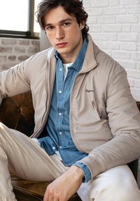 Ljus beige vadderad jacka över en blå denimskjorta. Modell sitter på en brun lädersoffa. Texturerad tegelvägg i bakgrunden.