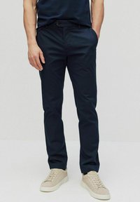 Donkerblauwe broek van soepele stof met zijzakken, een knoopsluiting, en een slim fit, gecombineerd met lichtbeige sneakers.