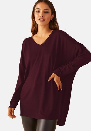 Frau trägt übergroßes dunkelviolettes V-Ausschnitt-Longsleeve mit schwarzen Lederleggings, posiert mit einer Hand in der Hüfte vor einfachem Hintergrund.