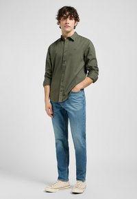 Olijfgroene overhemddres met lange mouwen, gecombineerd met lichtblauwe spijkerbroeken en beige sneakers; gladde stof, casual pasvorm en eenvoudig ontwerp.