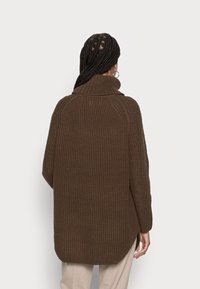 Weit gestrickter, dunkelbrauner Pullover mit hohem Kragen, überschnittenen Schultern und seitlichen Schlitzen. Strukturierter Stoff sorgt für ein gemütliches Aussehen.