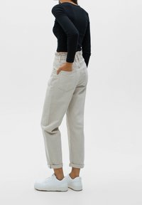 Personne portant un pantalon beige clair taille haute avec revers, un haut noir ajusté à manches longues et des baskets blanches, debout de profil avec une main dans la poche.