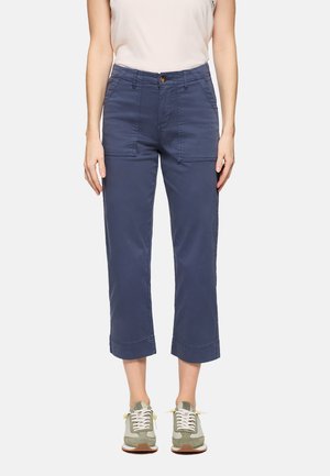 Mujer con pantalones cortos azul marino con bolsillos delanteros de parche, una blusa sin mangas blanca y zapatillas verdes y blancas, de pie contra un fondo blanco.