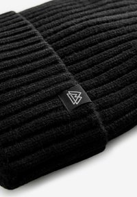 Bonnet noir en tricot avec texture côtelée et une petite étiquette noire avec un logo blanc sur le côté. Le matériau semble doux et épais.