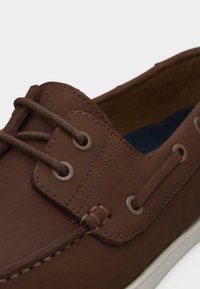Καφέ δερμάτινο παπούτσι τύπου boat με κορδόνια, λευκή σόλα, λεπτομερή ραφές και μεταλλικά ροζέτα.