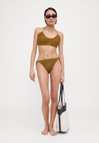 Khaki-colored bikini with textured fabric, halter top, and side-tie bottoms. Model holds a black and cream tote bag, standing barefoot.