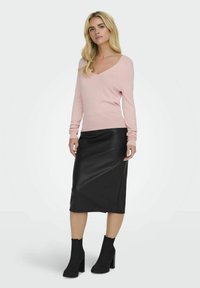 Helles rosa V-Ausschnitt-Pullover aus weichem Material, kombiniert mit einem schwarzen Midi-Rock aus Kunstleder und schwarzen Stiefeletten mit Blockabsatz.