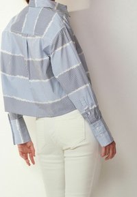 Chemise à rayures bleu et blanc avec col à pointes. Présente des manches longues avec boutons aux poignets et un pli sur la pièce d'épaule arrière. Portée avec un pantalon blanc.