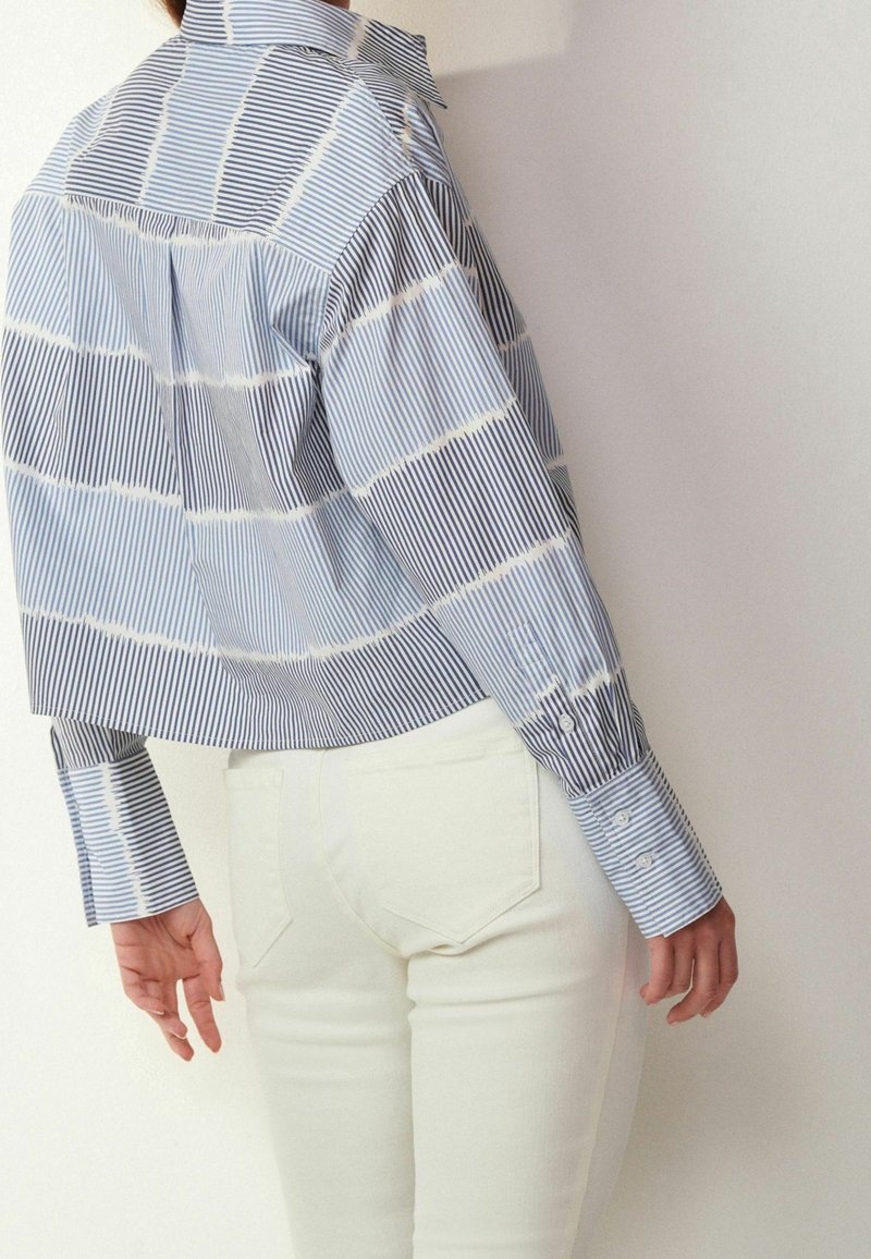 Chemise à rayures bleu et blanc avec col à pointes. Présente des manches longues avec boutons aux poignets et un pli sur la pièce d'épaule arrière. Portée avec un pantalon blanc.