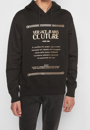 Czarna bluza z kapturem z złotym napisem „Versace Jeans Couture” i włoskim pismem, noszona z ciemnymi dżinsami przez stojącą osobę na jednolitym tle.