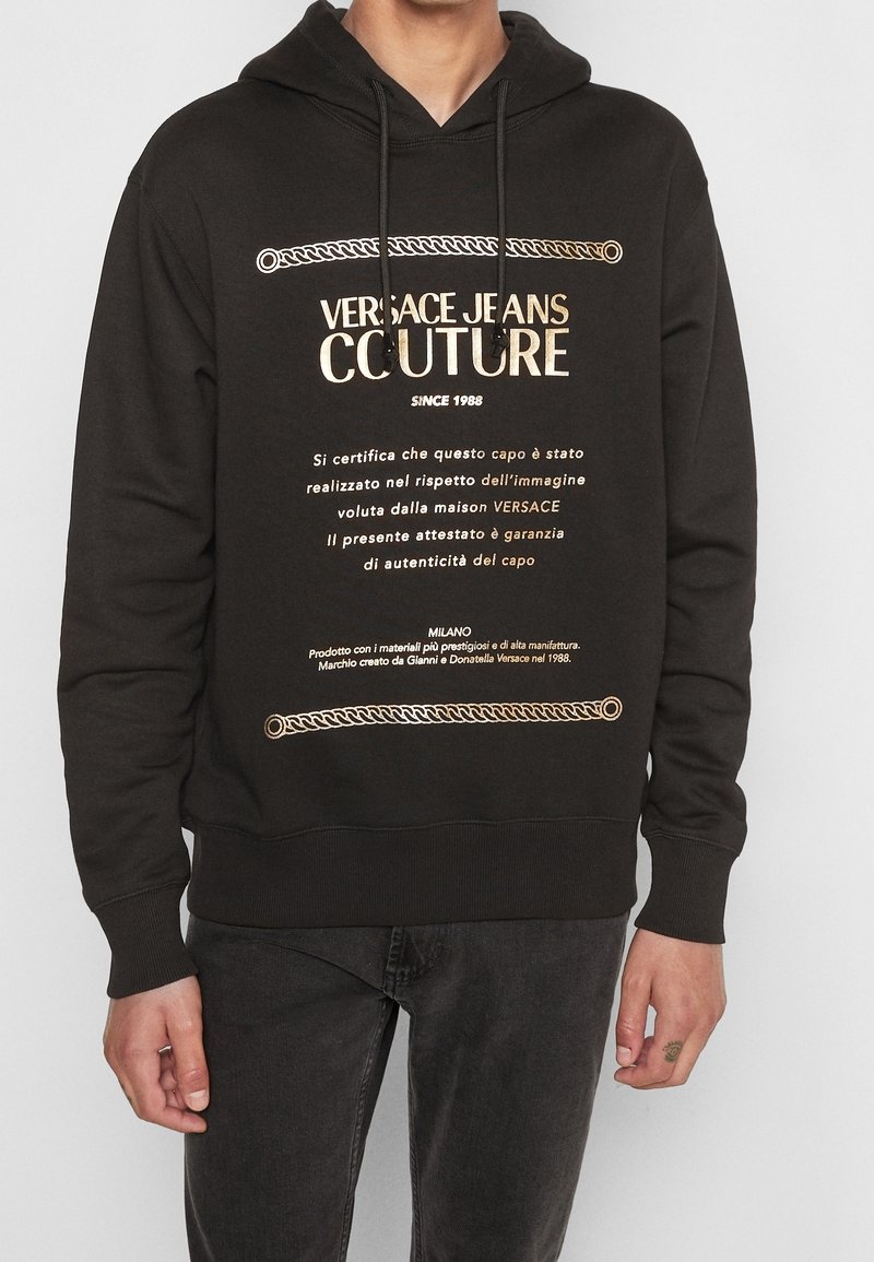 Sweat à capuche noir avec inscription dorée "Versace Jeans Couture" et écriture italienne, porté avec un jean foncé par une personne debout sur un fond uni.