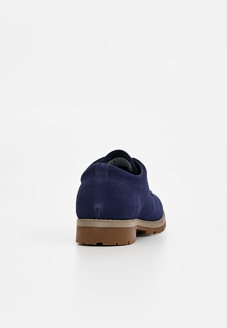 Scarpa in suede blu navy con suola in gomma marrone, vista da dietro su uno sfondo bianco uniforme.