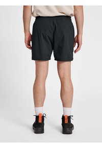 Czarne szorty z elastycznym pasem, bocznymi kieszeniami i gładką teksturą. Widok z tyłu pokazuje osobę w czarnych butach z pomarańczowymi akcentami.