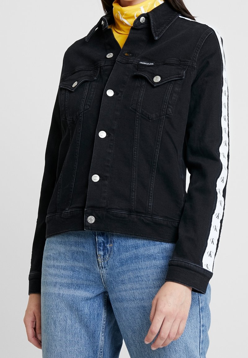 Black denim jacket with snap buttons, two front pockets, and white logo accents down the sleeves. Features a classic collar and fitted design.