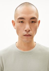 Hellgrüner, strukturiertes Strickcrewneck-Sweatshirt. Nahaufnahme einer Person mit glatze und neutralem Ausdruck, die sich auf die Stoffdetails konzentriert.