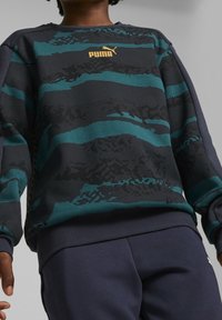 Felpa blu scuro con motivi a onda in teal e nero, con polsini e orlo a costine, caratterizzata da un logo Puma dorato sul petto. Texture morbida.