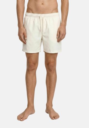 Homme debout pieds nus portant un short élastique uni écru avec cordon, sur un fond clair.