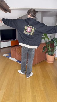 Črna oversized kapuca z grafiko, ki prikazuje rdečo in zlato grb na hrbtu, nosi jo preko sivih kavbojk in belihtenisic.