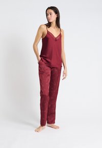 Camisole en satin bordeaux avec bordure en dentelle, associée à un pantalon assorti présentant un délicat motif floral. Le mannequin est pieds nus devant un fond uni.