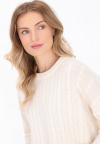 Maglione crema a maglia intrecciata con scollatura rotonda, motivo testurizzato e tessuto morbido. Accompagna onde morbide nei capelli e trucco minimale.