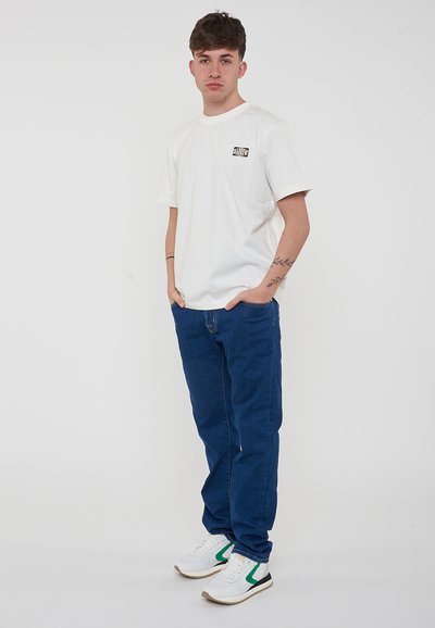 Camiseta blanca de algodón con un pequeño logo negro en el pecho izquierdo, combinada con pantalones de mezclilla azules y zapatillas blancas con acentos verdes.