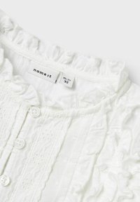 Camicia bianca in cotone con righe verticali, scollatura arricciata e quattro bottoni. L'etichetta riporta "name it" con i dettagli della taglia.
