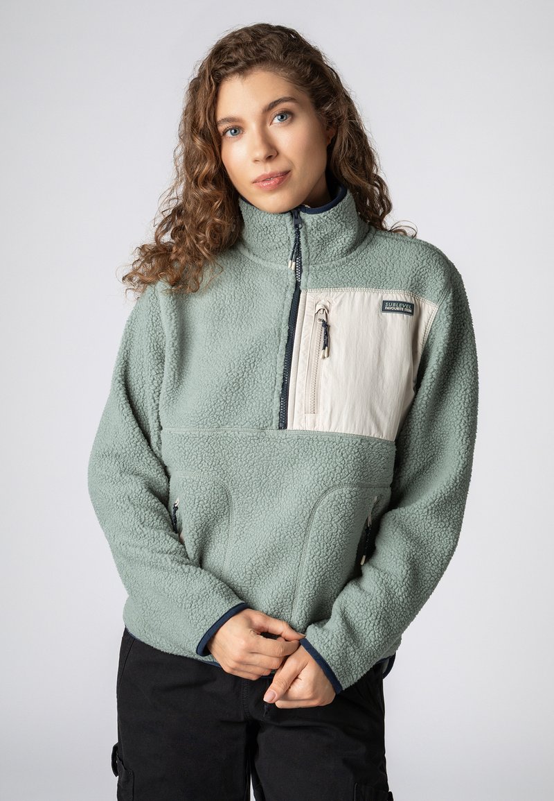 Fleece pullover in pastelgroen met een hoge kraag, zijzakken en een crèmekleurige zakpaneel. Voorzien van een ritssluiting en marineblauwe accenten.