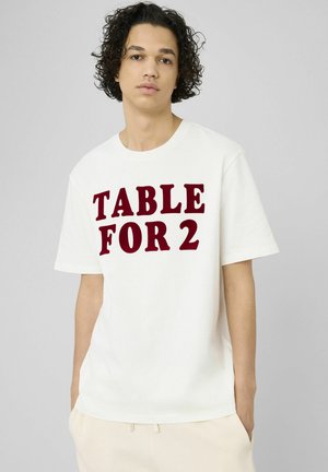 Jeune personne aux cheveux bouclés portant un T-shirt blanc avec un grand texte rouge "TABLE FOR 2", debout les mains dans les poches sur fond gris.