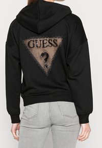 Sweat à capuche noir avec un dos en maille triangulaire, texte "GUESS ?" en sequins noirs et surpiqûres décoratives. Tissu doux, coupe décontractée.
