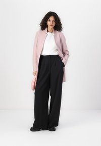 Cappotto rosa chiaro con taglio dritto, indossato sopra un top bianco. Pantaloni neri a gamba larga, abbinati a una piccola borsa rosa. Texture morbide.