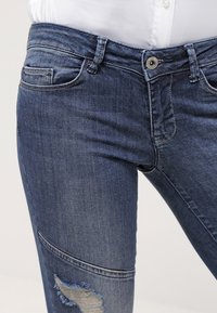 Jeans de mezclilla azul con un diseño ajustado, que cuentan con una zona de rodilla desgastada, bolsillos frontales y un cierre de botón en la cintura.