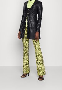 Giubbotto di pelle nera con bottoni dorati, abbinato a pantaloni a zampa d'elefante in stampa leopardata gialla brillante. Sandali con tacco alto neri e lucidi completano l'outfit.